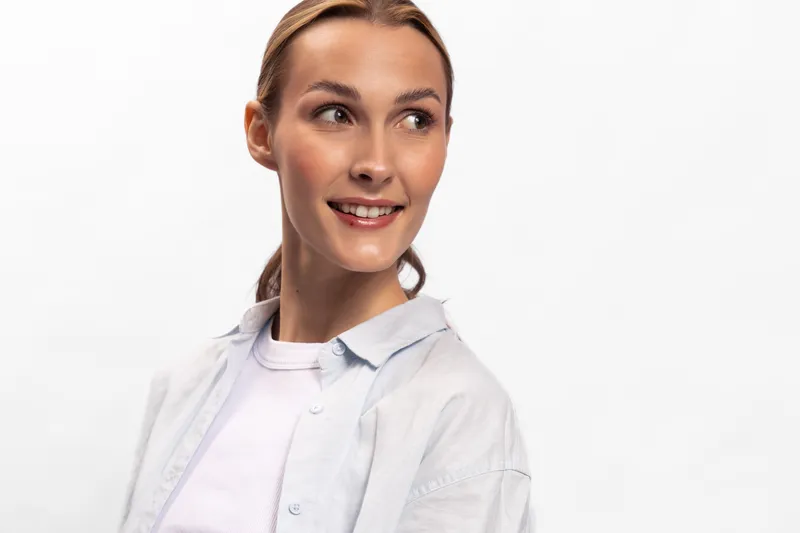 Smiling young woman with blonde hair looks sideways, wearing white shirt and shirt jacket. Bright studio lighting highlights natural skin and positive expression on white background.