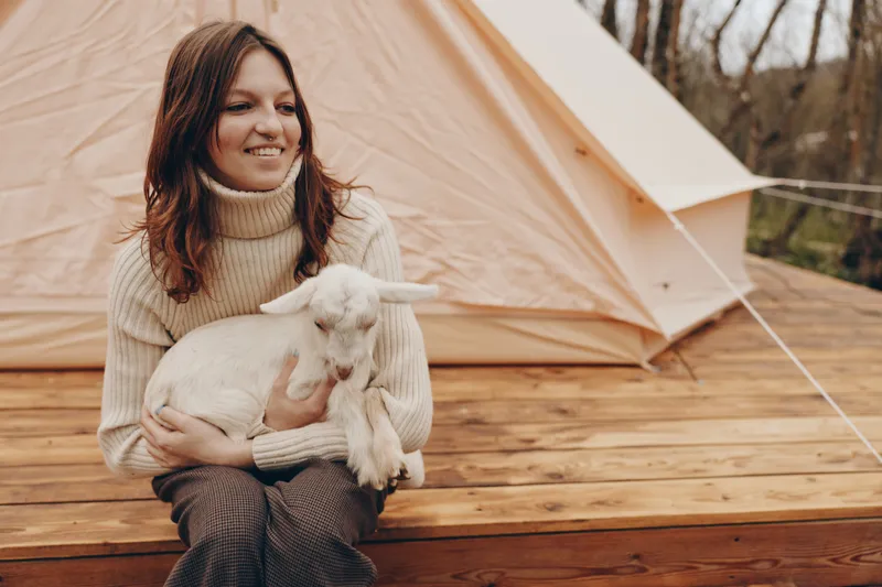 Young woman with brunette hair holds a white baby goat while sitting on wooden steps near a beige tent in an autumn outdoor setting with a cozy lifestyle mood.