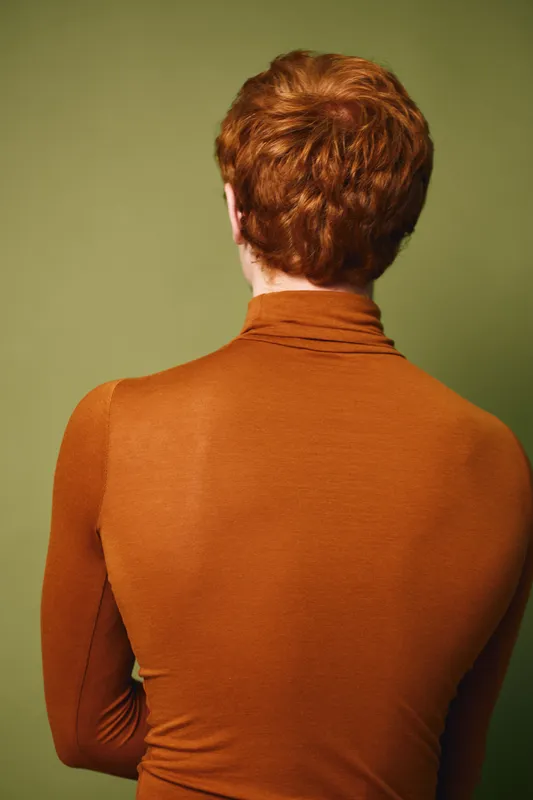 Person with red hair wearing brown turtleneck sweater standing with back to camera on green background. Portrait style, casual clothing and studio setting.