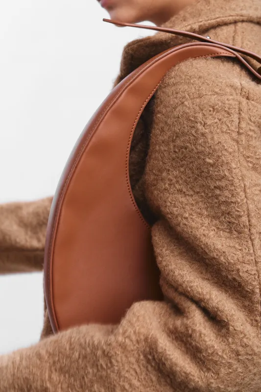 asian woman wearing a cozy camel coat and a leather backpack, seen from the side in soft light, conveying warmth and style in a modern urban setting.