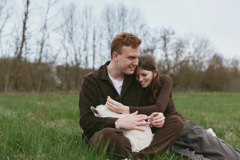 A couple sits on lush grass in an open field, cuddling a goat kid, capturing a calm outdoor lifestyle moment in nature, highlighting affection, companionship, and relaxed rural happiness.