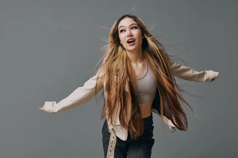 Young woman with long hair wearing casual clothes is dancing and posing with arms outstretched, happy expression on face in studio with gray background.