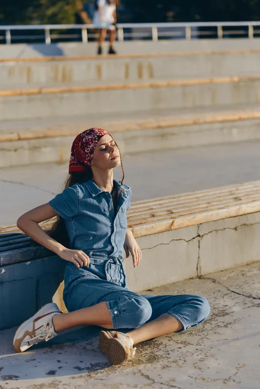 Lifestyle woman in modern boho-western denim street style relaxing outdoors with film color effect, casual outfit, red bandana, and natural sunlight creating a warm atmosphere.