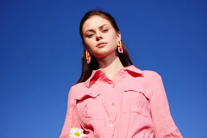 Young woman in a pink shirt and colorful earrings, standing against a bright blue sky, holding a daisy, expressing confidence and joy in a vibrant outdoor setting