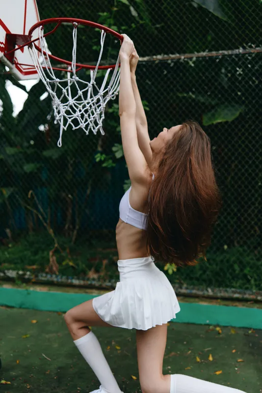 Young athletic girl wearing a sports outfit, performing a slam dunk on a basketball court surrounded by greenery, showcasing strength and determination