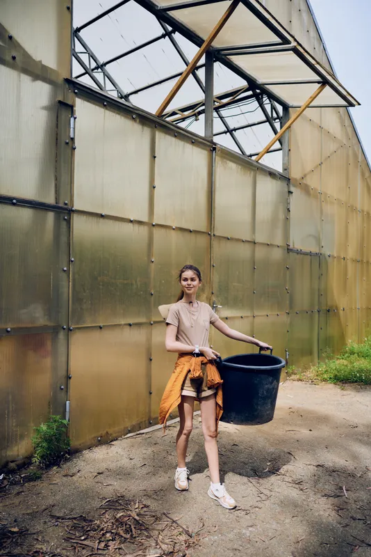 Girl woman at greenhouse carrying black bucket for gardening and agriculture work on a farm outdoors in casual summer clothes, young female gardener walking near plant house.