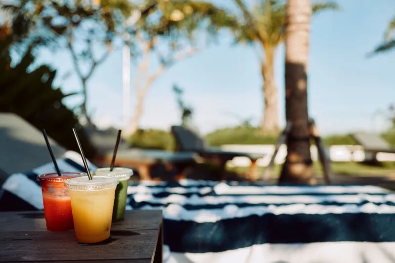 Relaxed outdoor poolside scene with colorful drinks on a wooden table, clear water and palm trees in the background, conveying a warm summer mood, casual leisure, and sunny relaxation. style. Fun.