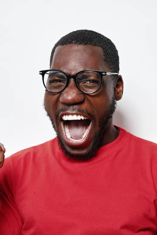 African American man with dark skin wearing glasses and red shirt shouting with excitement and clenched fist on white background. Emotion, expression and celebration concept.