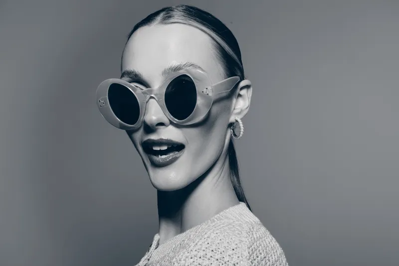 Young Caucasian woman with pale skin wears large round sunglasses and knitted sweater posing with open mouth. Studio portrait in black and white lighting.