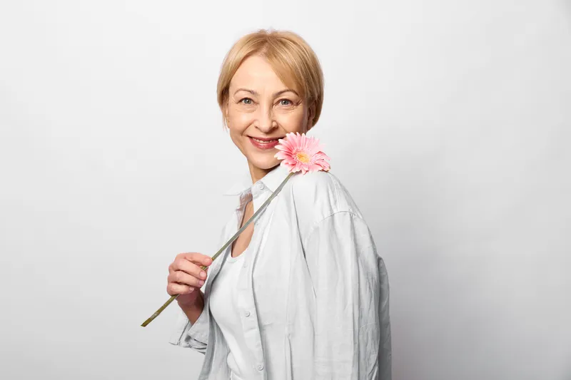 Woman holding a pink flower, studio closeup portrait of elderly person in white coat, gentle smile, soft light, tidy hair, calm mood, bright neutral background