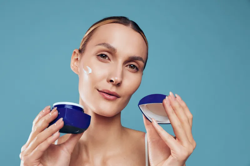 A young woman with clear skin applies cream to her face holding a blue jar and lid against a plain blue background. Concept of skincare, beauty routine, and natural complexion.