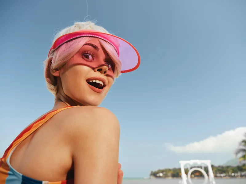 A cheerful woman in a colorful swimsuit and pink visor smiles toward the camera on a sunny beach, with a clear sky and distant waves in the background.