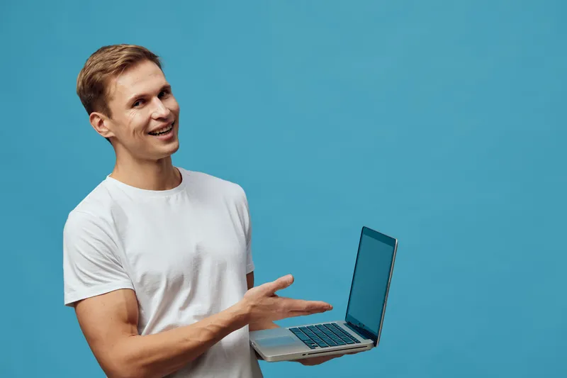 Smiling young caucasian man in casual white t-shirt holding open laptop computer, gesturing with hand, isolated on solid blue background, studio portrait, people lifestyle concept, technology, communi