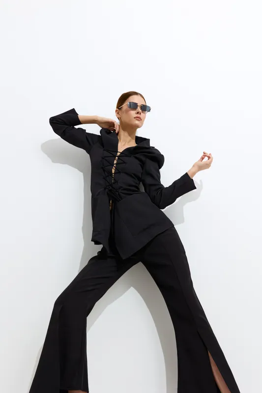 Fashion woman in stylish black outfit posing confidently against white wall, wearing sunglasses with modern, edgy vibe and high slit trousers, expressing bold elegance.