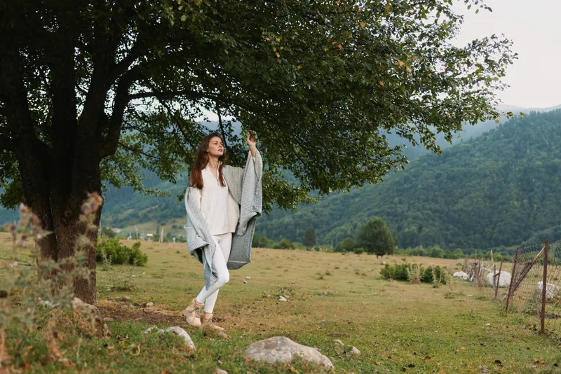 Young woman with long brown hair walks through a meadow beneath a large tree, wearing a light cardigan and white sweater, enjoying a carefree outdoor moment on a sunny, breezy day.