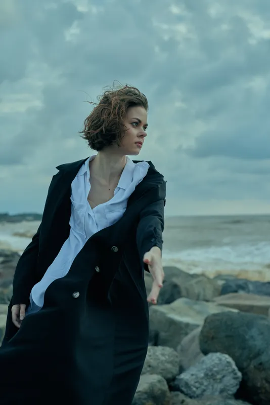Woman on a rocky shoreline facing strong wind in a long coat and white shirt, moody ocean backdrop and cloudy sky, dynamic pose conveying drama, fashion photography in outdoor setting