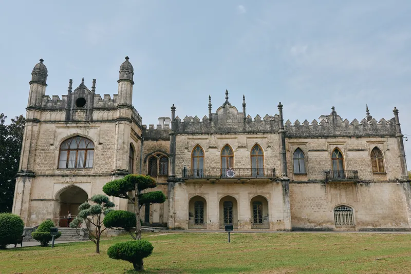 Majestic historic castle with ornate stone facade and tall arches rises over a manicured lawn with trimmed trees under a clear sky, evoking timeless architectural grandeur.