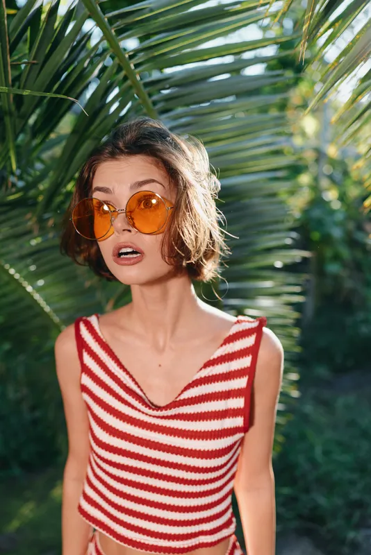 A young woman in a red and white striped tank top and orange round sunglasses stands outdoors among tropical greenery, her expression surprised and playful, bright sunlight framing her face.
