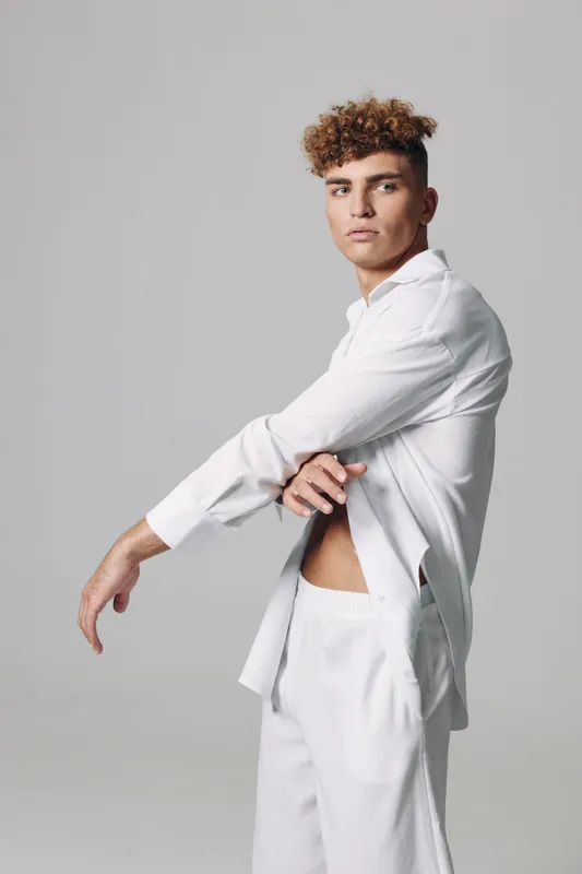 Young man with curly hair wearing white shirt and pants posing in studio with neutral background. Fashion model showing casual style and confident expression in minimalistic setting.