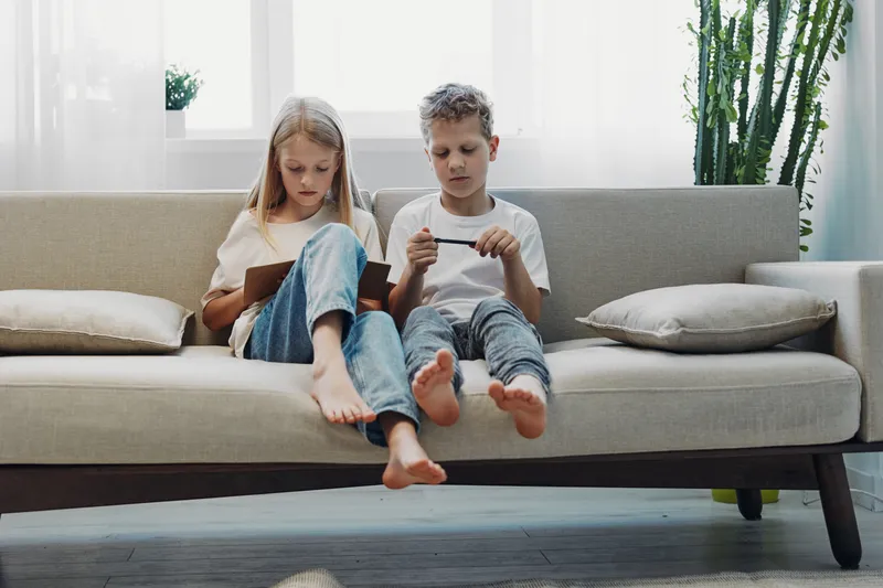 Two barefoot children sitting on a modern sofa focused on digital devices in a bright living room with natural light and green plants.