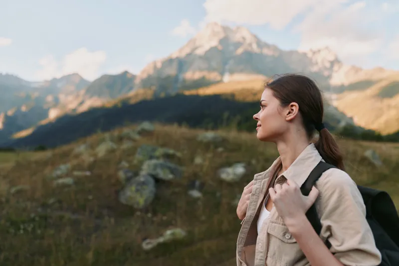 A young brunette woman with a backpack gazes toward distant mountains from a hillside, wearing a calm, curious expression as the wind brushes her jacket and grass sways in golden alpine light.