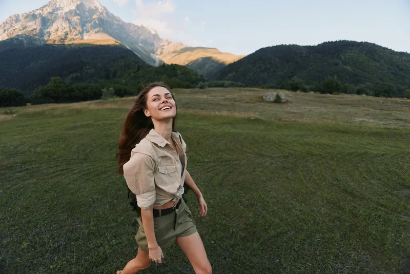 Young woman enjoying outdoor lifestyle, smiling in a grassy field with mountain landscape, wearing a casual beige shirt and shorts, basking in sunny natural scenery.