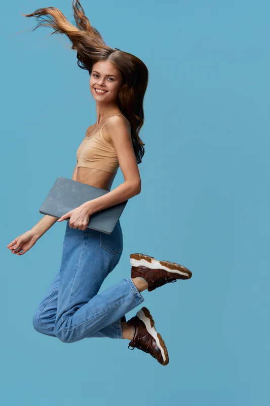 young woman jumping with laptop, casual blue jeans and beige top, smiling, isolated on blue background, joyful energy and motion captured vividly