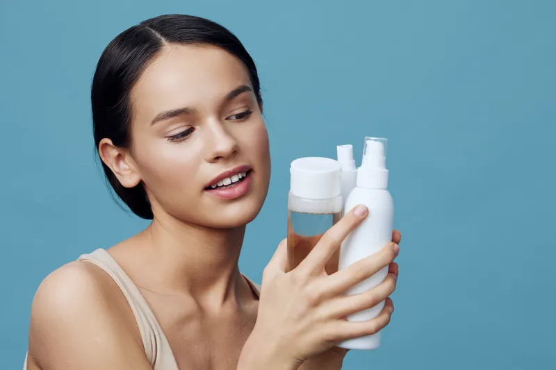 Young woman holding skincare products, showcasing beauty and self care with a serene expression against a soft blue background, emphasizing healthy skin and beauty routine