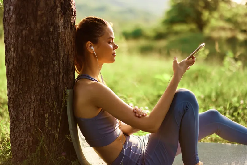 Young woman in blue activewear, enjoying music on her phone while sitting by a tree in a sunny outdoor setting, conveying relaxation and wellness