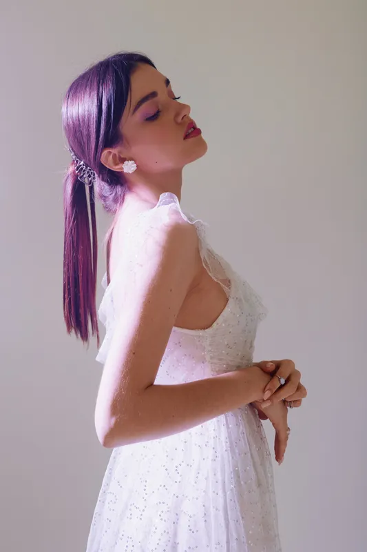 Young woman in a white dress poses with eyes closed and head tilted back. Soft lighting highlights her beauty and fashion style. Concept of elegance and confidence.