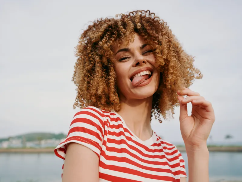 Energetic portrait of a cheerful young woman with curly hair wearing a red and white striped shirt, smiling at the camera against a bright outdoor backdrop.