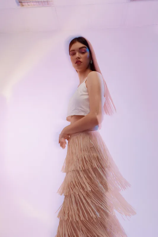 Young woman in a white top and layered fringe skirt poses confidently in a soft pastel studio. Concept of woman beauty fashion with elegant and modern style.