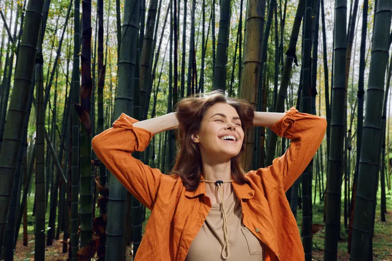 Woman in orange jacket smile in bamboo forest, nature portrait of a relaxed woman outdoors with hands behind head, joyful expression and peaceful lifestyle wellbeing in greenery.