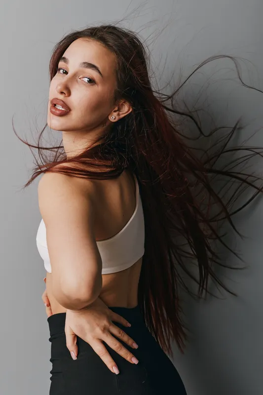 Young woman with long hair wearing white top and black pants posing against gray background. Hairstyle flowing and natural makeup with confident expression and casual fashion style.