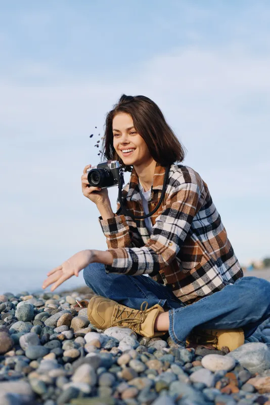 Photographer, woman, camera, beach, rocks, outdoor, casual, smiling while capturing scenes on a sunny shoreline with lens in focus and candid coastal mood