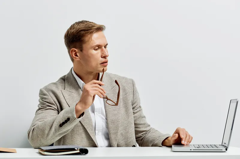 Professional young man in smart casual attire working at desk with laptop and glasses, holding pen, focused on screen in modern office environment, business and technology concept.
