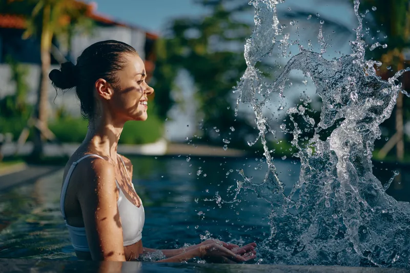 A smiling woman enjoys a refreshing moment at an outdoor pool, water spraying as a fountain arcs nearby. A lifestyle mood shot focusing on leisure, relaxation, and sunny summer recreation.