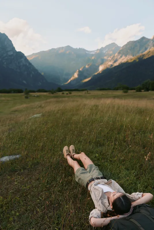 Woman relaxing in a grassy field near mountains, enjoying outdoor life, casual outfit, lying on her back with arms behind her head, serene natural scenery and peaceful mood