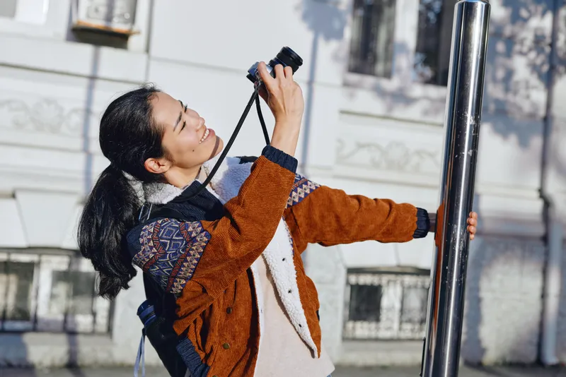 Woman photographs outdoors on an urban street with a camera, capturing candid moments and architectural details in daylight, showcasing photography workflow and street style