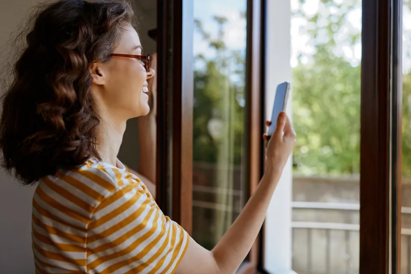 Woman lifestyle home, smiling woman by a window holds a smartphone, casual striped shirt, warm daylight, relaxed moment of everyday connection in a cozy living space.
