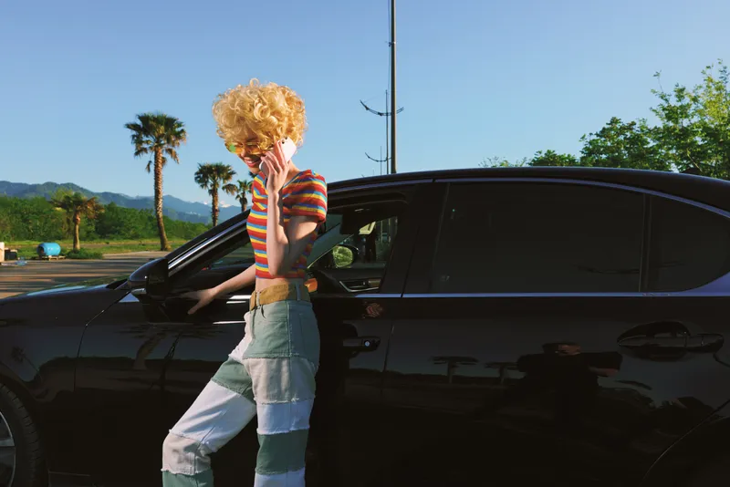 Woman exits a sleek black car in a retro striped top, stylish jeans, and curly hair, posing on a sunny street with palm trees, conveying casual lifestyle and vintage film color.