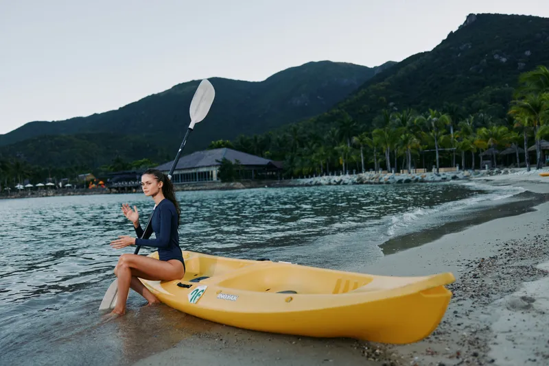 A kayaker sits on a bright yellow kayak at a calm tropical beach, preparing to paddle as gentle waves kiss the shore and distant mountains frame the scene.
