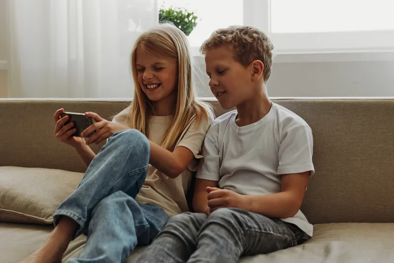 Two happy children sitting on sofa smiling and watching smartphone screen together in cozy living room environment, casual home leisure moment.