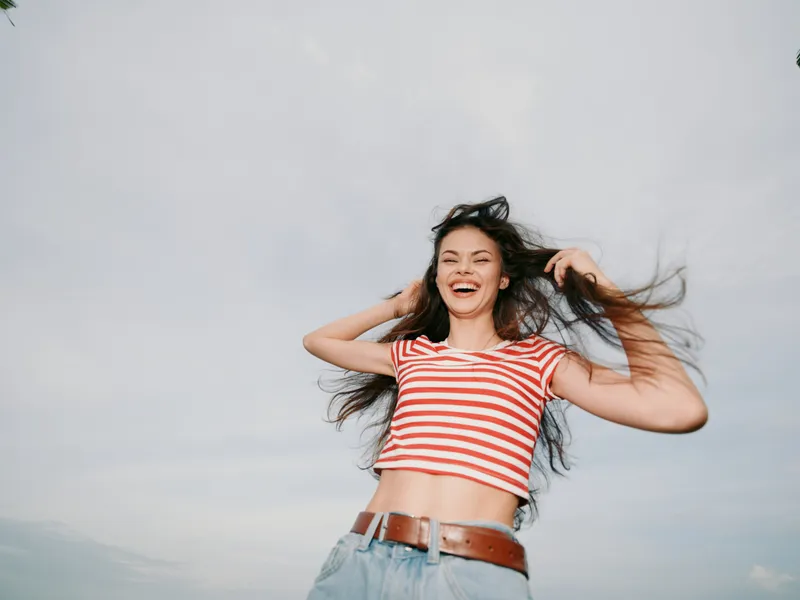 Young woman joyfully spins outdoors, wearing a red and white striped crop top and denim jeans, capturing carefree summer energy and a bright, uplifting mood.