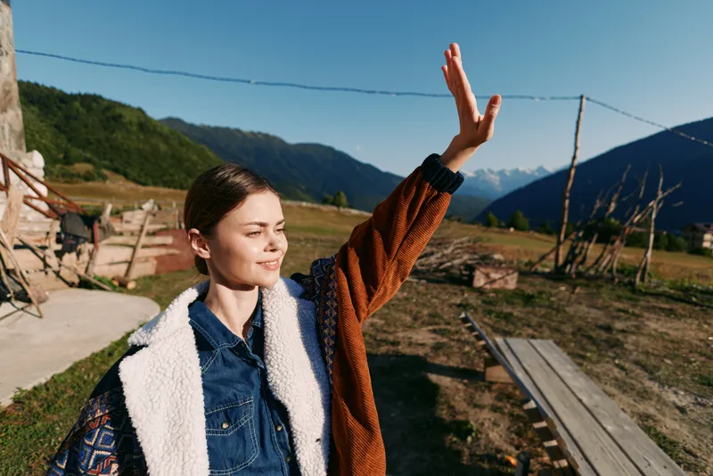 Woman smiling in the mountains, waving hand outdoors in the countryside, wearing a denim jacket with shearling lining in a portrait-style shot on a sunny rural meadow.