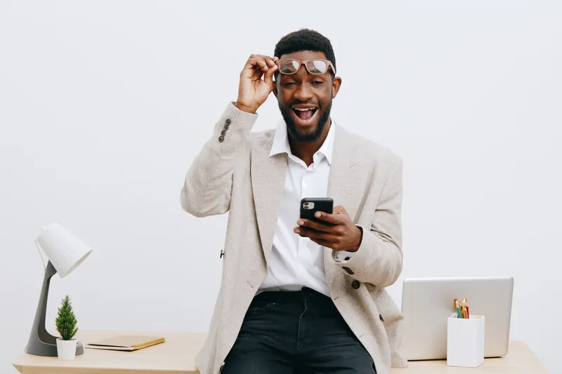 Smiling young man with glasses holding mobile phone and looking excited in modern office setting with minimalistic desk and accessories, casual business attire, positive emotions, lifestyle concept.
