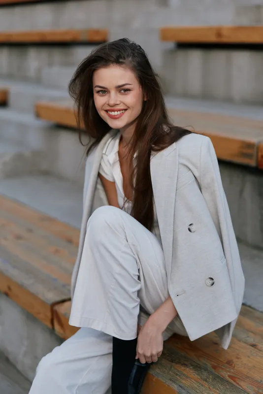 Confident business woman in stylish light outfit sitting on wooden bleachers, showcasing modern professionalism with a bright smile and relaxed pose.