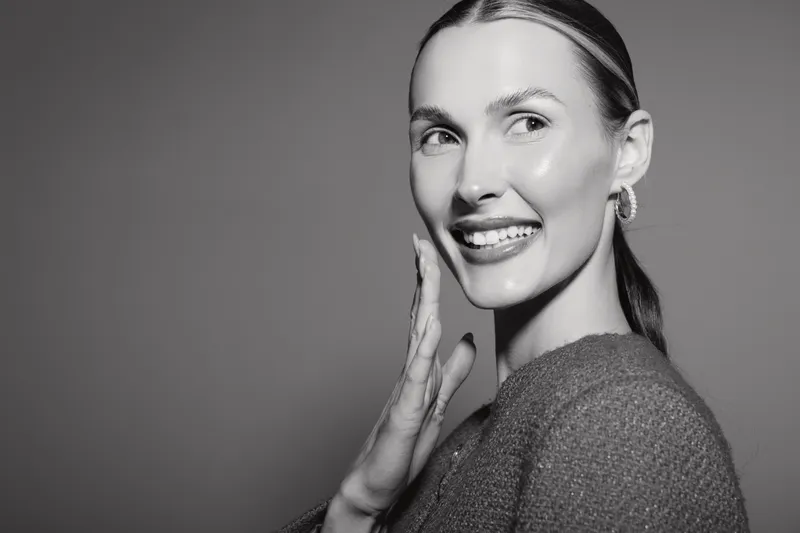 Smiling Caucasian woman with light skin touches her face gently in close-up portrait. Concept of skincare, beauty, health, and natural glow with soft studio lighting on grey background.