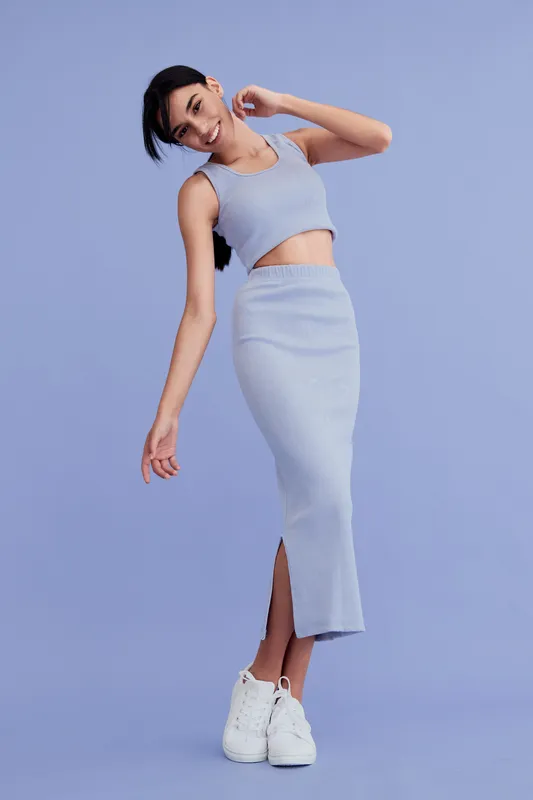 Smiling woman in a stylish light blue dress and white sneakers poses gracefully against a plain soft purple background, radiating joy and modern fashion sense.