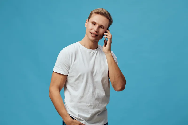 Young smiling man in white t-shirt talking on smartphone standing on blue background casual style digital nomad communication concept